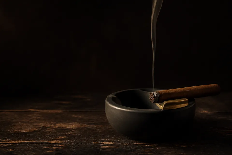 Cendrier en céramique noire au repose-cigare laiton, un cigare allumé y déroule un mince filet de fumée